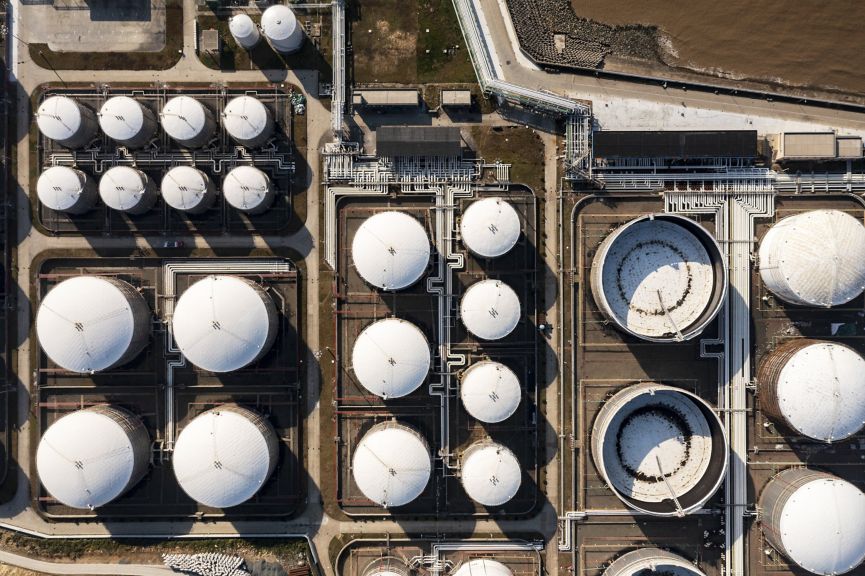 A top-down aerial view displays a complex industrial site featuring numerous large circular storage tanks connected by a network of pipelines.