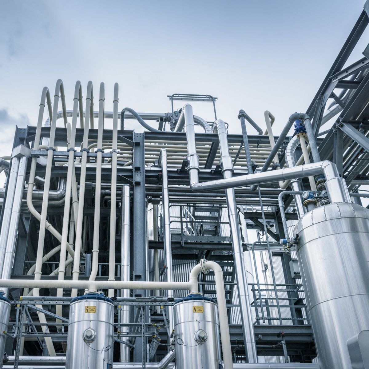 A low-angle view of complex industrial pipes and stainless steel processing tanks against a cloudy sky.