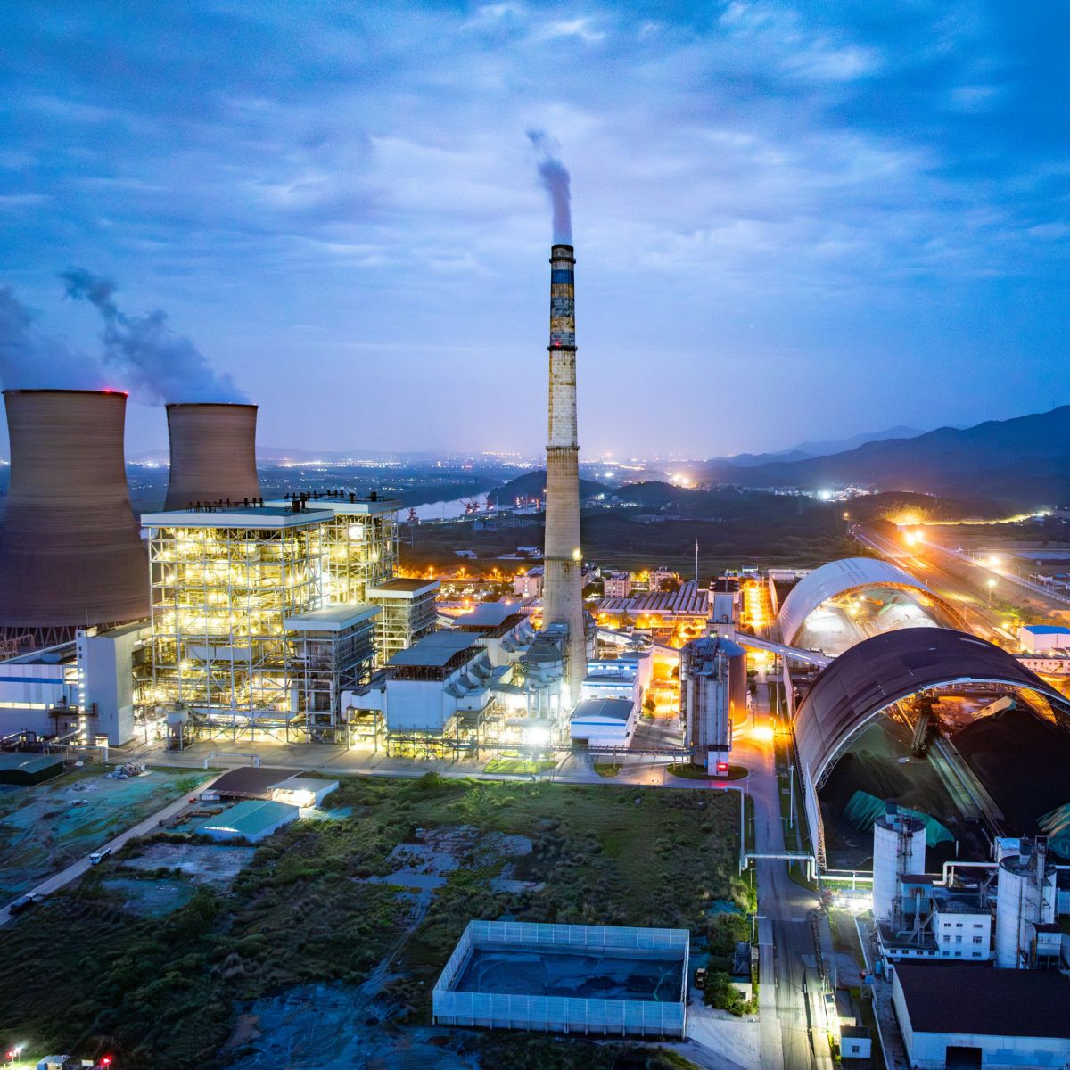 An aerial view captures a thermal power plant at twilight, featuring illuminated infrastructure, cooling towers, and large coal storage domes.