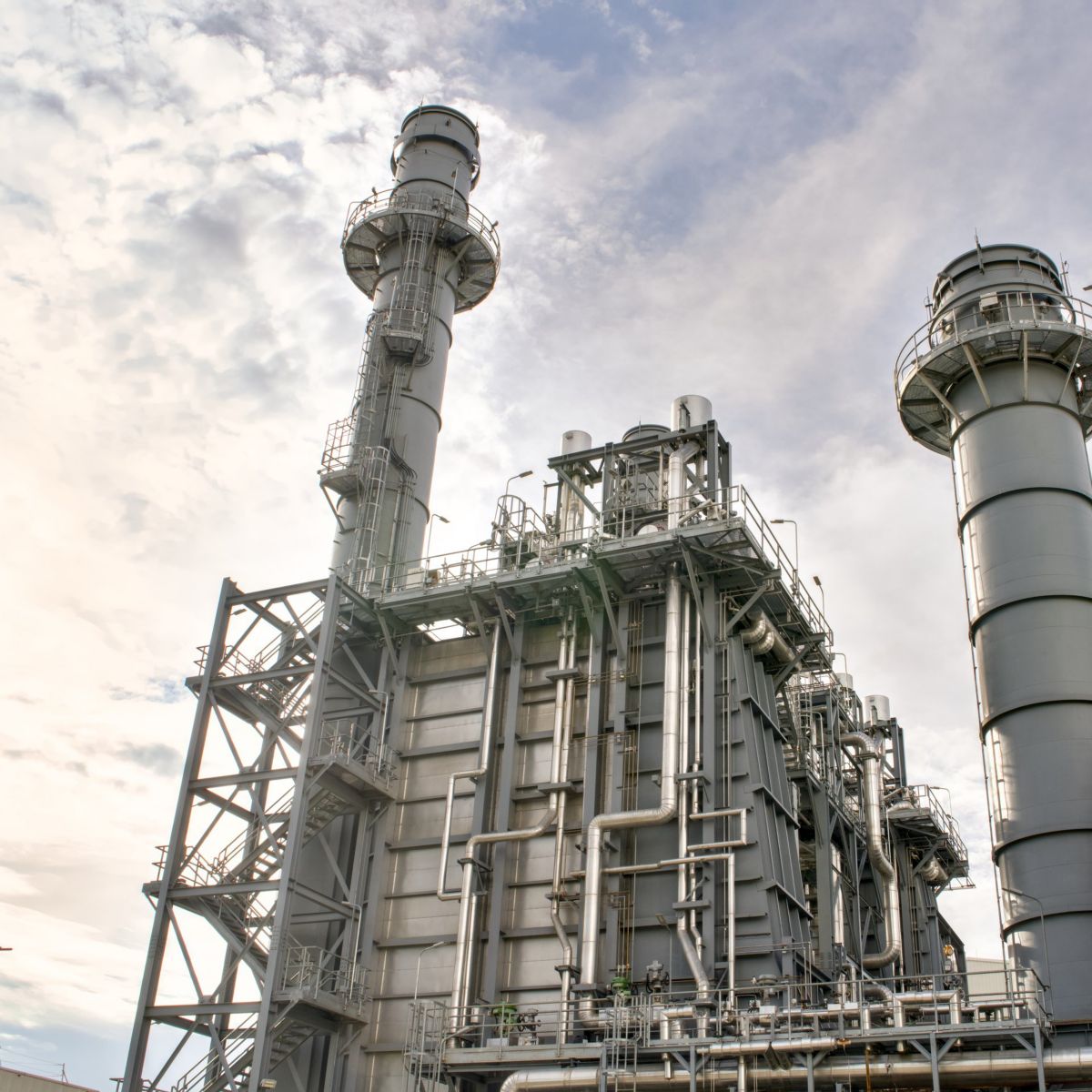 A low-angle shot of industrial power plant structures featuring steel exhaust stacks and complex piping against a cloudy sky.