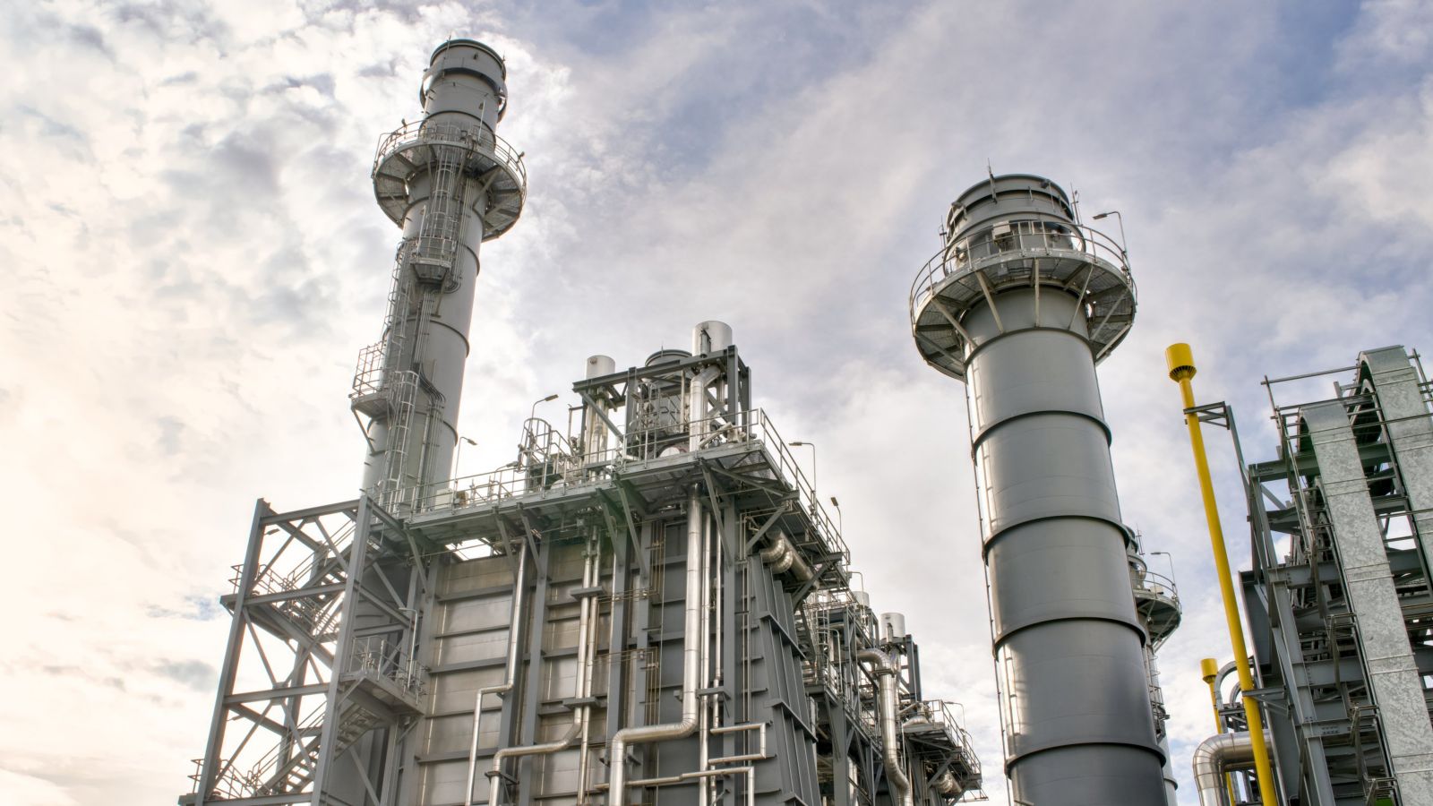 A low-angle shot of industrial power plant structures featuring steel exhaust stacks and complex piping against a cloudy sky.