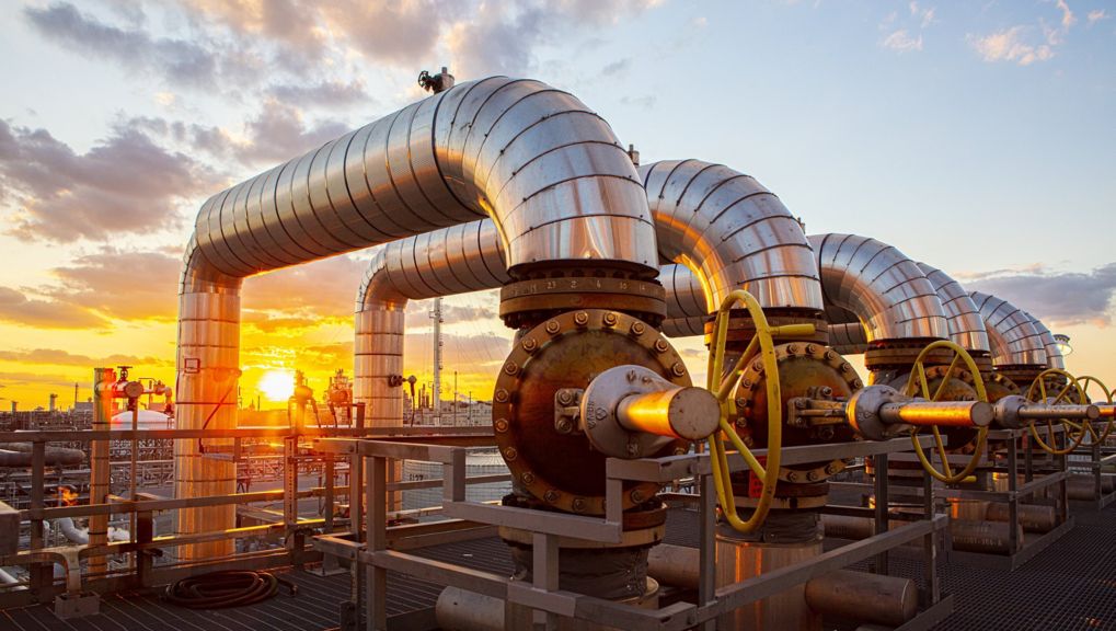 Industrial metal piping and manual control valves backlit by a golden hour sunset at a refinery facility.