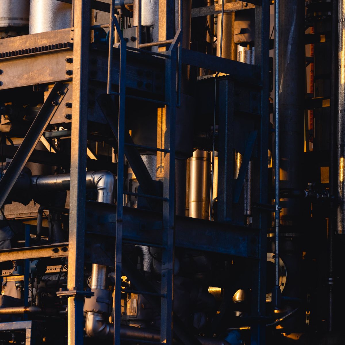 Sunlight illuminates metallic pipes and structural steel frames within a dense industrial refinery complex at dusk.