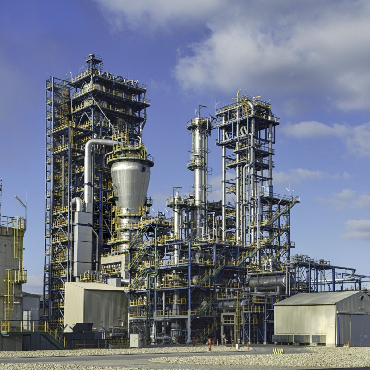 Wide exterior view of an industrial refinery plant with towers, piping systems, and support structures under a cloudy sky.
