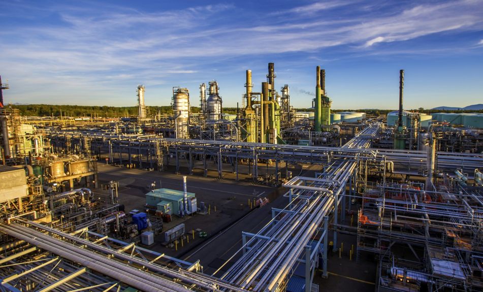 A wide-angle view of a sprawling industrial refinery complex with extensive piping systems and towering processing units under a clear blue sky.