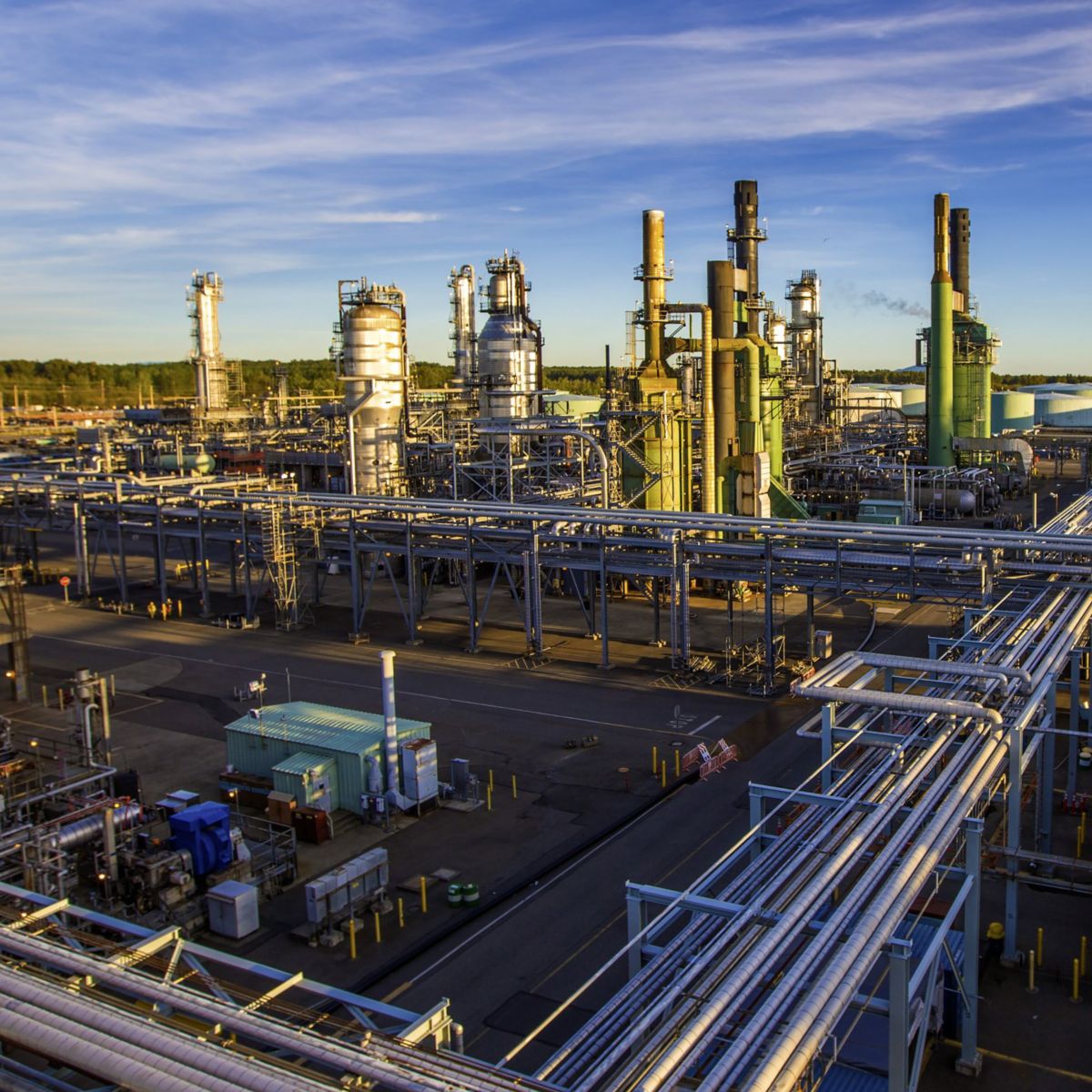 A wide-angle view of a sprawling industrial refinery complex with extensive piping systems and towering processing units under a clear blue sky.