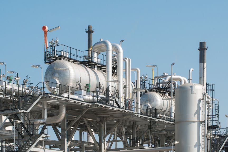 A detailed view of high-pressure storage tanks, complex piping systems, and steel gantries within an industrial oil refinery against a clear blue sky.