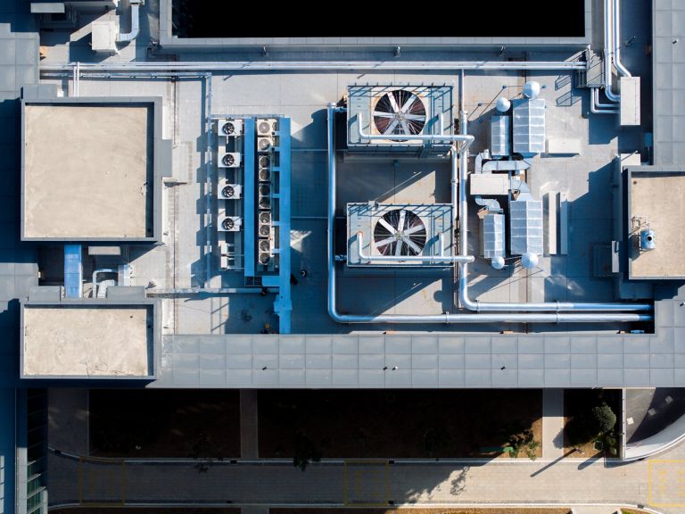 An aerial view displays large industrial ventilation units and piping arrays on a flat commercial building rooftop under bright sunlight.