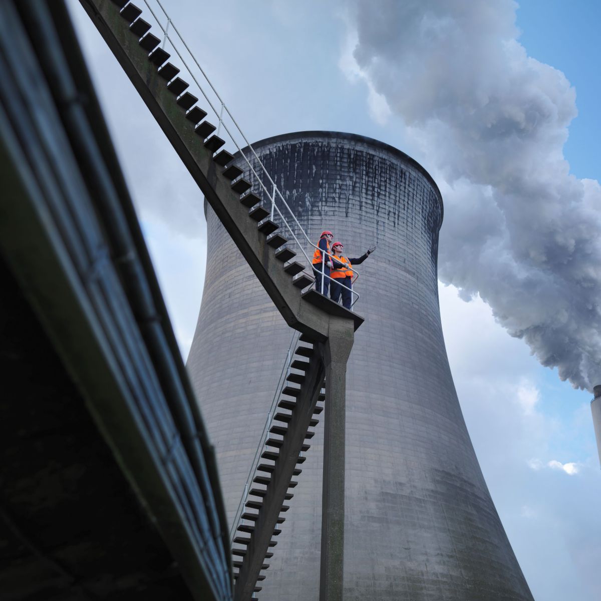 Two workers in safety gear assess emissions near cooling towers, showcasing industrial safety practices.