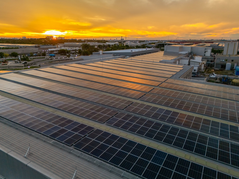 Aerial view of a vast rooftop solar panel installation illuminated by a vibrant, golden sunset over an industrial manufacturing park.