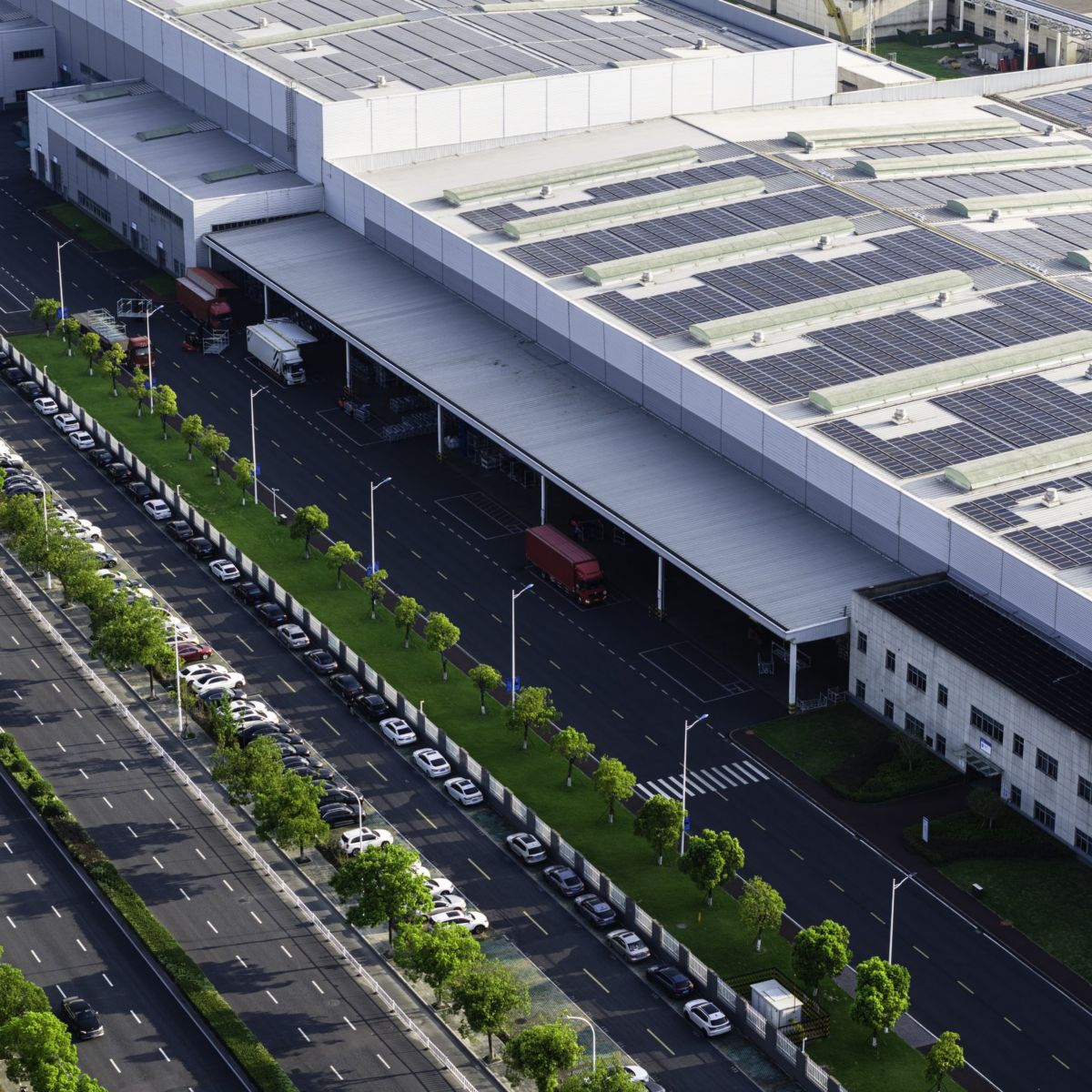 An aerial view of a large warehouse complex featuring extensive roof-mounted solar panels and a busy parking and loading area.