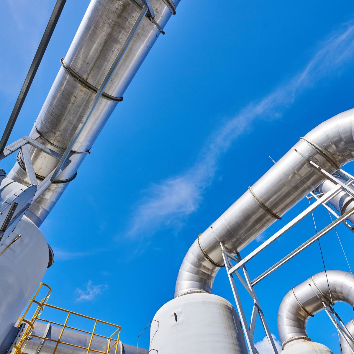 A low-angle view captures complex metal piping and industrial infrastructure against a bright, clear blue sky.