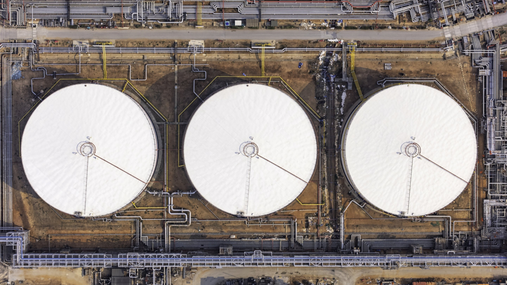 An aerial view captures three large white industrial storage tanks surrounded by an extensive network of connecting pipelines within a facility.