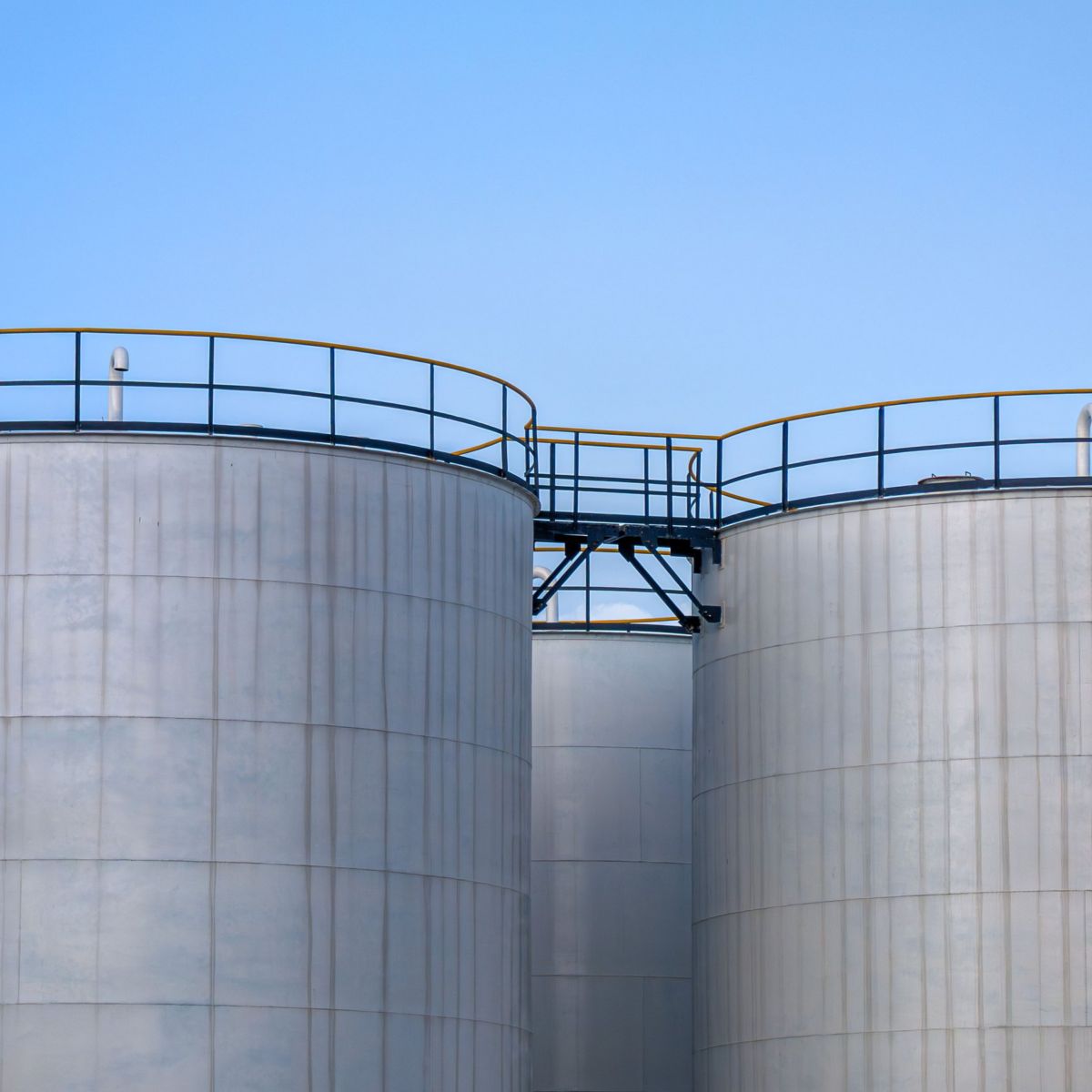 Large white cylindrical industrial storage tanks interconnected by a metal catwalk against a clear blue sky.