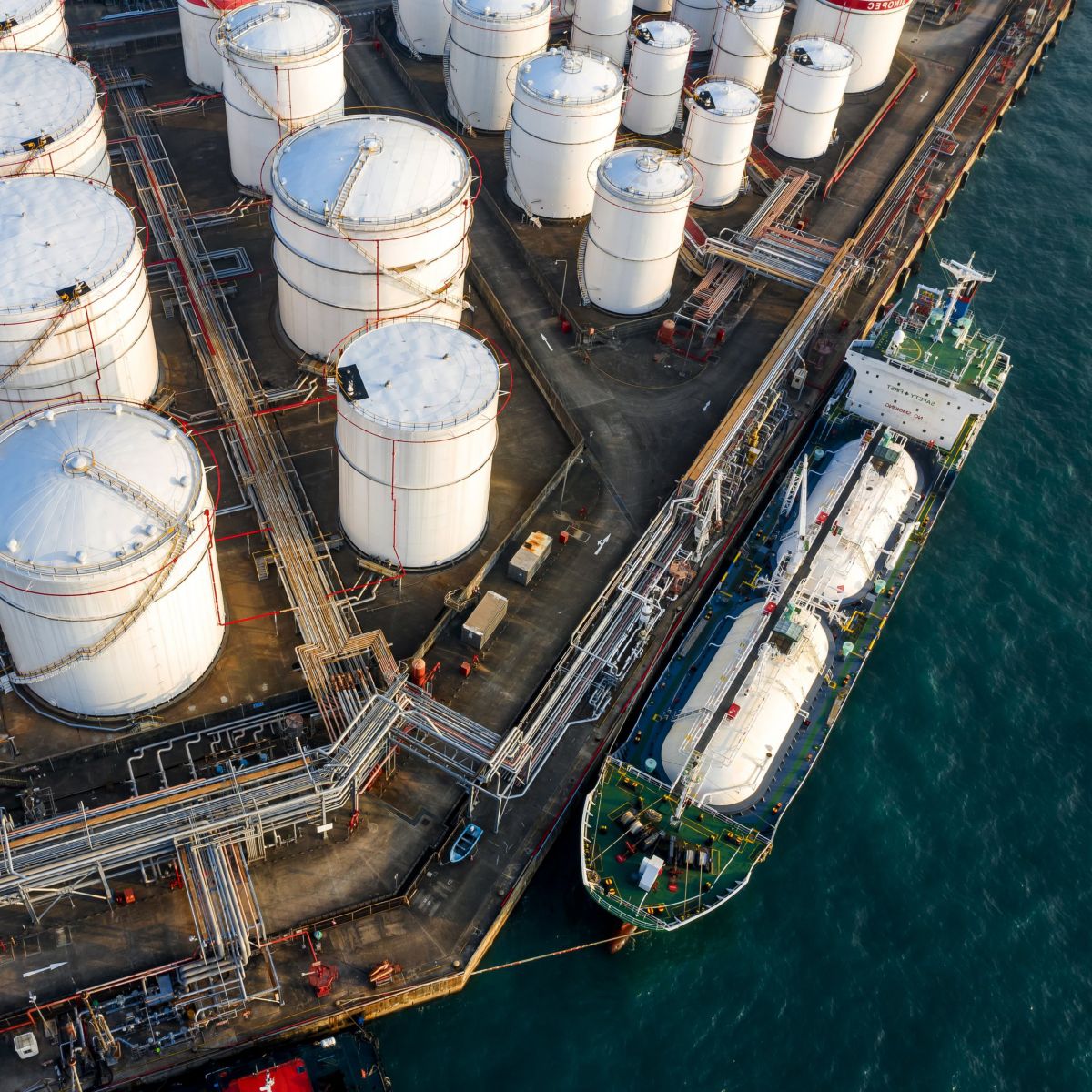 Aerial view of an industrial terminal featuring white storage tanks and a tanker ship docked at a loading pier.