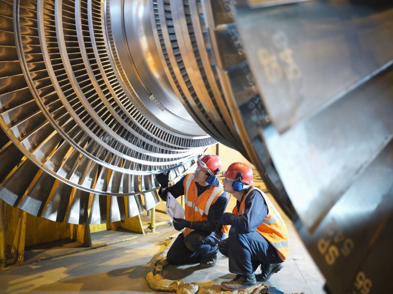 Technicians wearing safety gear inspect a large power generation turbine rotor in an industrial facility.