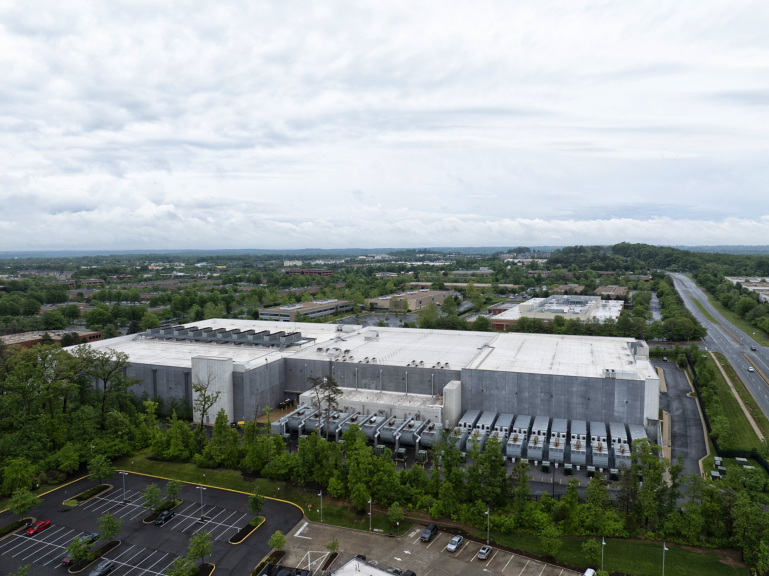Aerial view of a large industrial facility featuring expansive rooftop cooling units and surrounding parking areas under an overcast sky.