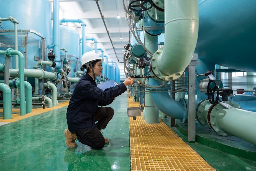 A female technician in protective gear inspects industrial piping and valves inside a modern water treatment facility.