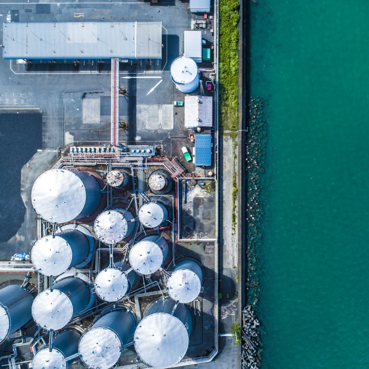 An overhead view captures industrial fuel storage tanks situated directly along the edge of a vibrant teal waterway.
