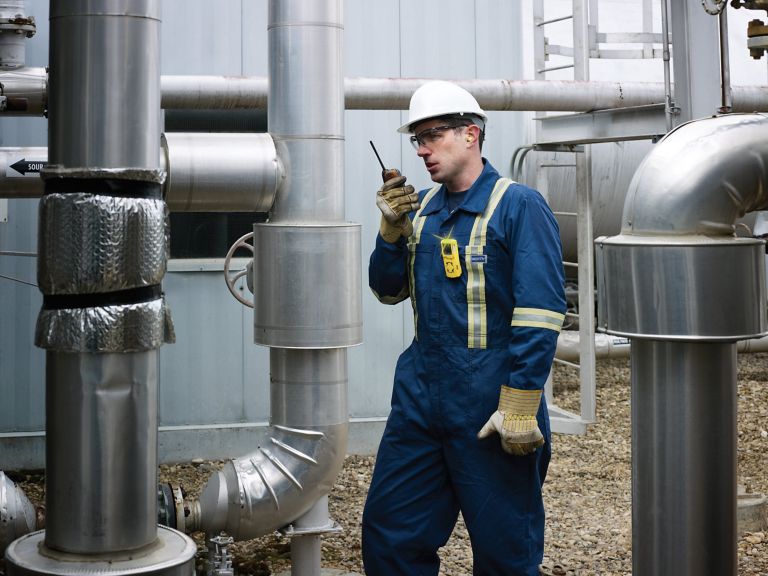 An industrial operator in safety gear communicates via two-way radio while inspecting piping at a refinery facility.