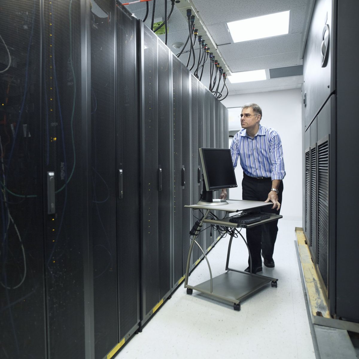 A technician moves a computer workstation through a data center aisle alongside rows of server racks.