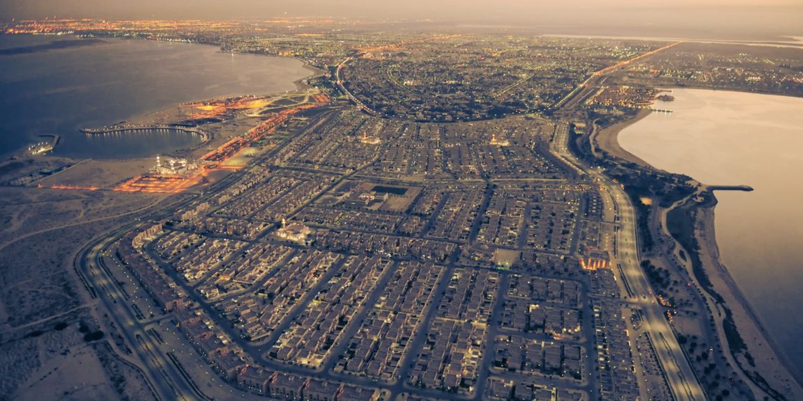 Aerial night view of a coastal city with illuminated roads, waterfront areas, and dense residential development.
