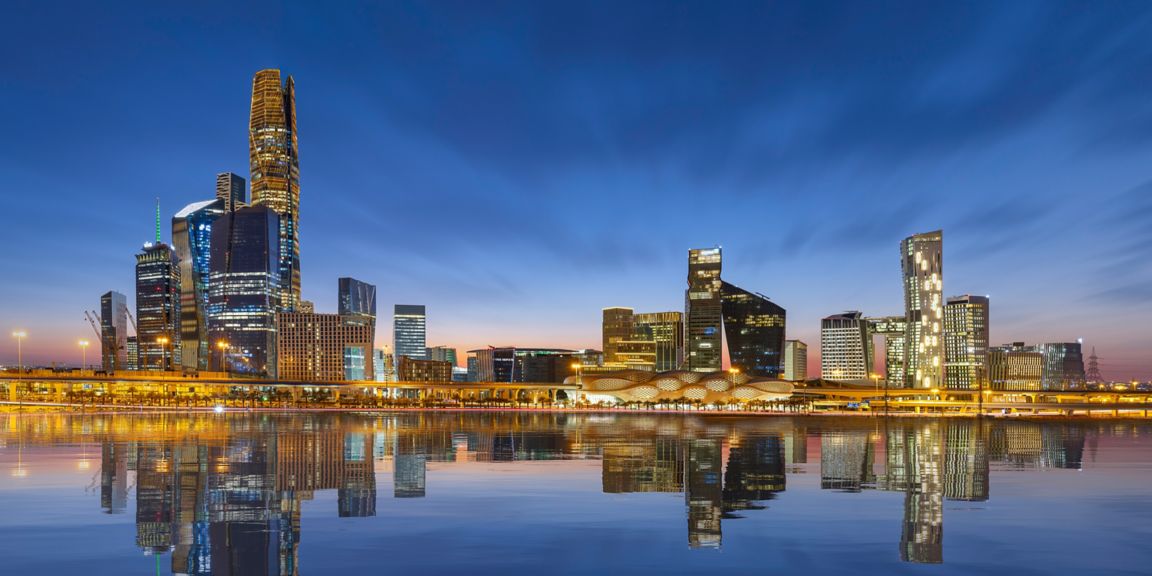 City skyline with high-rise buildings reflected in calm water, featuring illuminated architecture in an urban environment.
