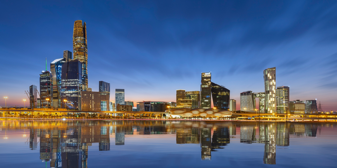 City skyline with high-rise buildings reflected in calm water, featuring illuminated architecture in an urban environment.