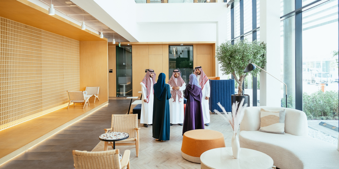 Group standing and discussing in a modern office lounge area with seating, plants, and natural light from large windows.