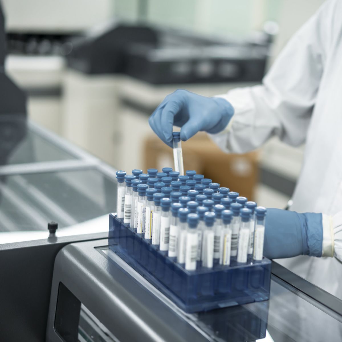 A technician in a lab coat and protective gloves processes a rack of test tubes in a modern clinical diagnostic laboratory.