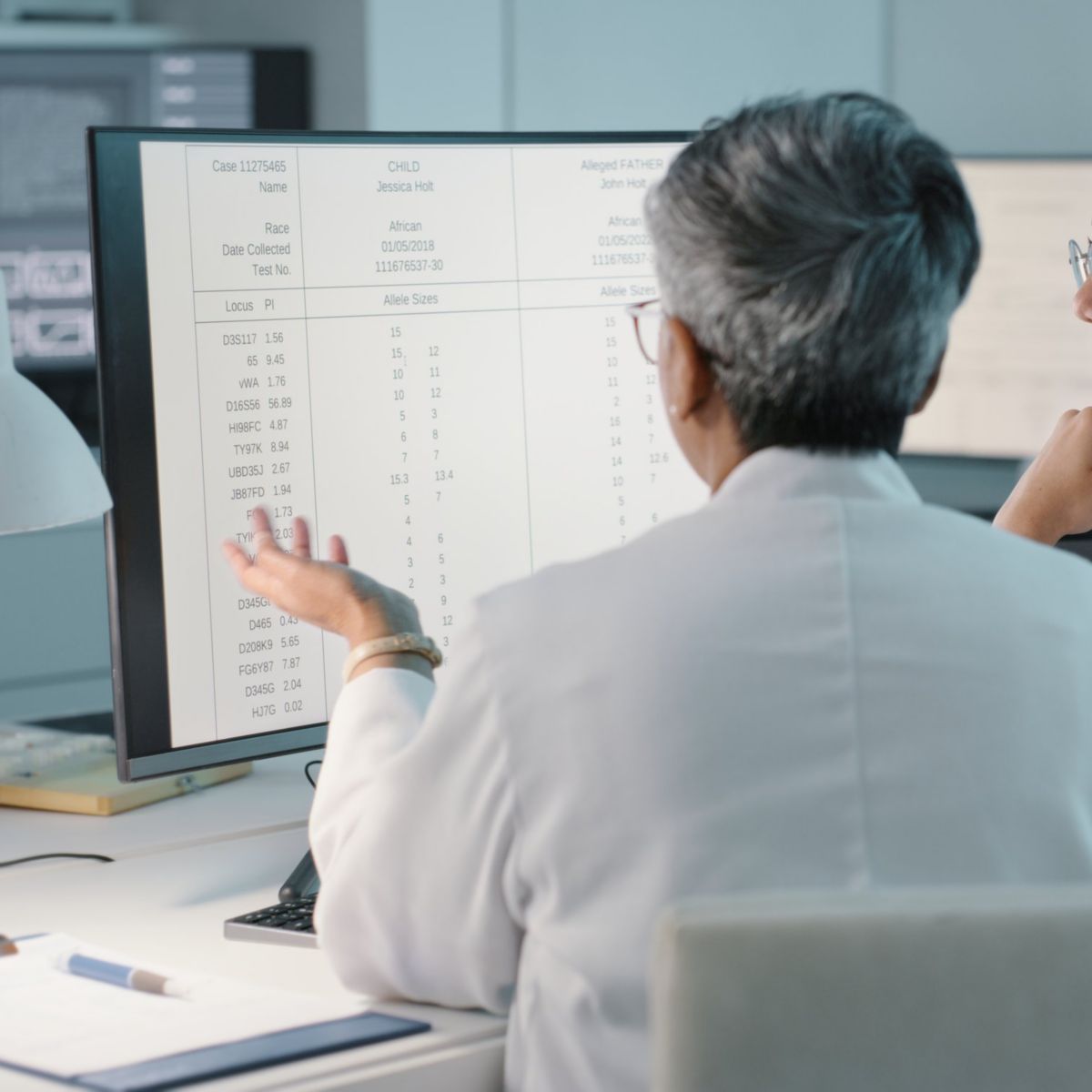 Two scientists in lab coats examine digital DNA test results on a computer screen within a professional medical research facility.