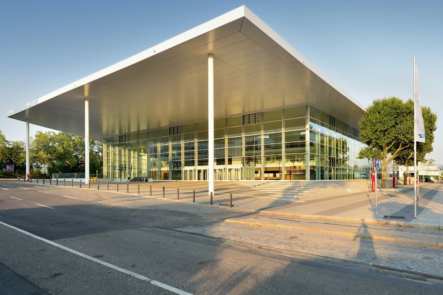 Modern glass-fronted exhibition hall at Messe Essen with a large cantilevered roof, wide entrance plaza, and surrounding walkways, serving as a venue for trade shows and industry events.