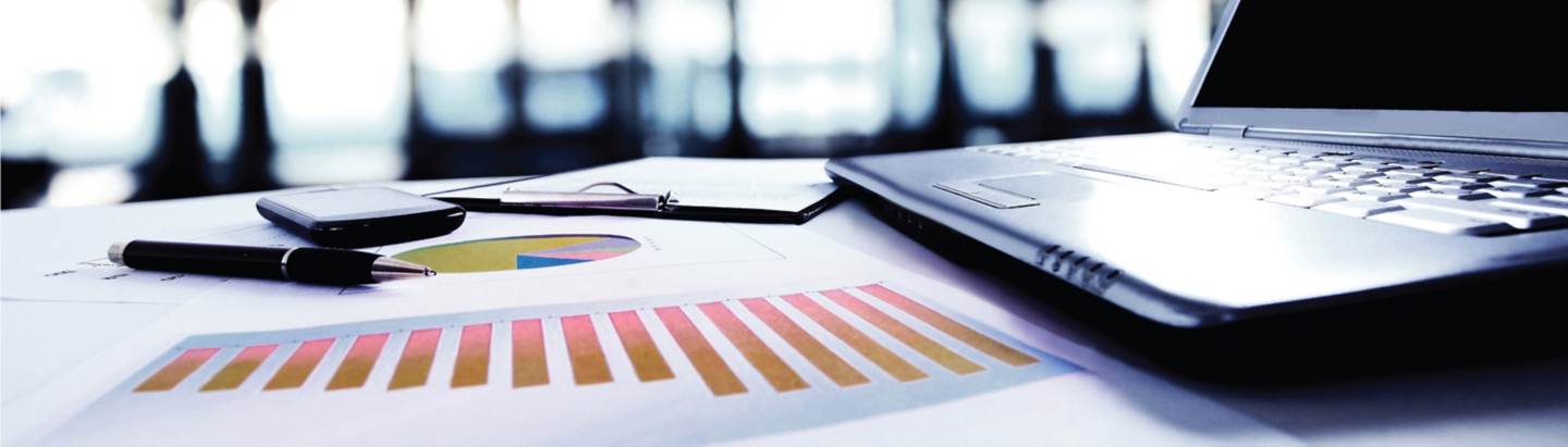 Laptop placed on a desk beside printed charts with colored bar graphs in a workspace setting with a blurred background.