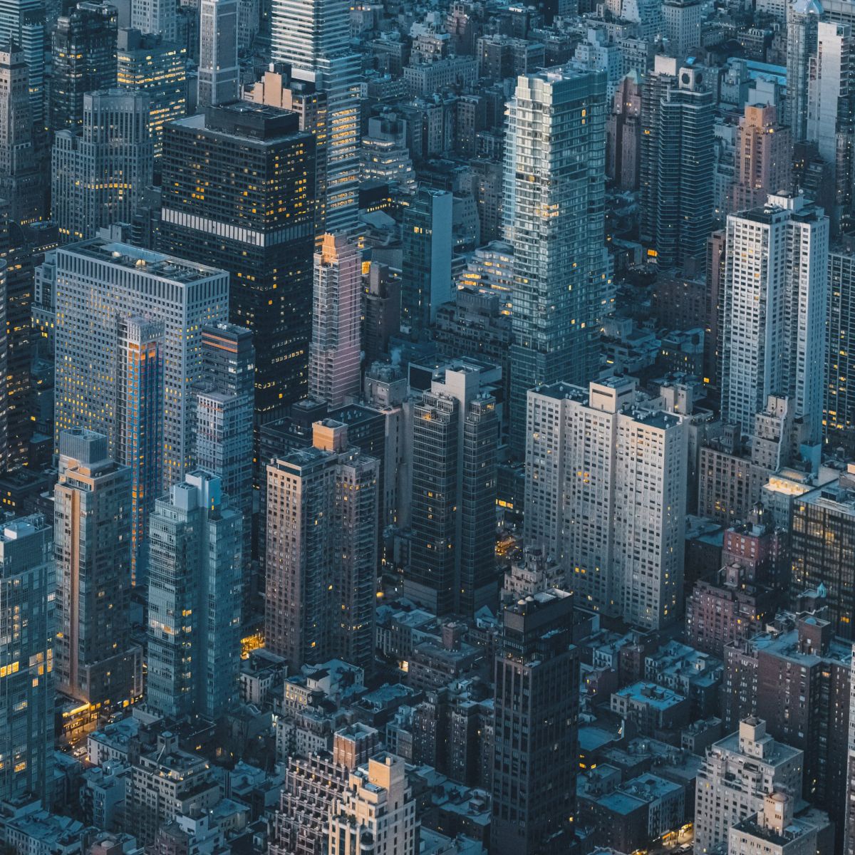 An aerial view of dense Manhattan skyscrapers captured during twilight with warm interior lighting illuminating the dark city grid.
