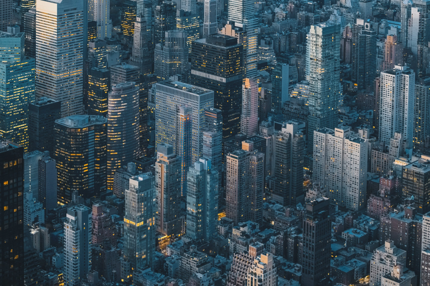 An aerial view of dense Manhattan skyscrapers captured during twilight with warm interior lighting illuminating the dark city grid.