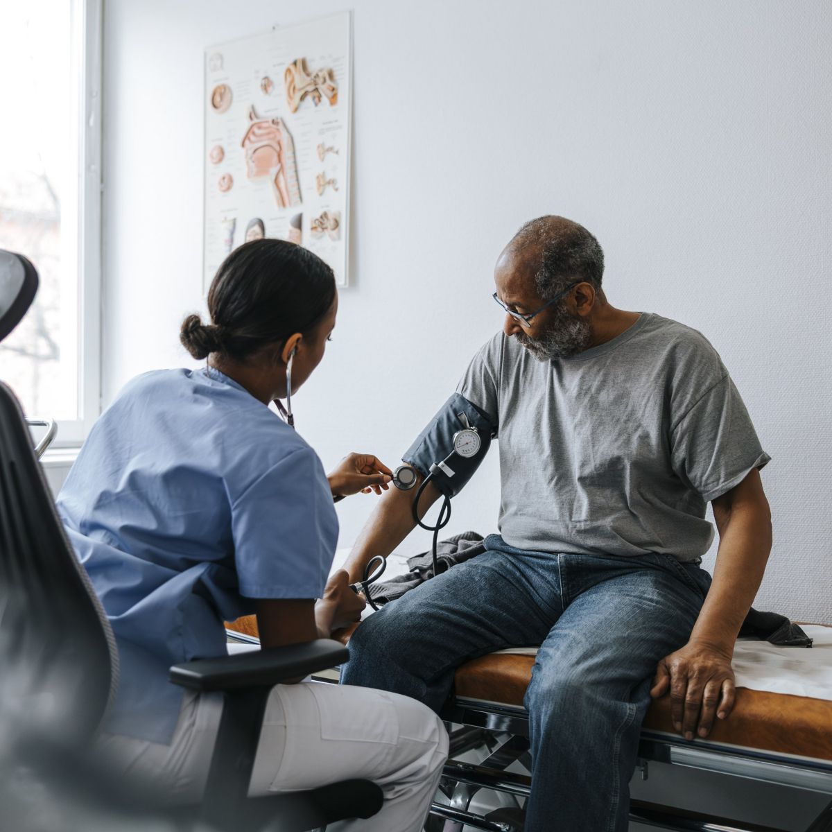 A female nurse uses a stethoscope and cuff to measure the blood pressure of an elderly male patient in a clinical office setting.