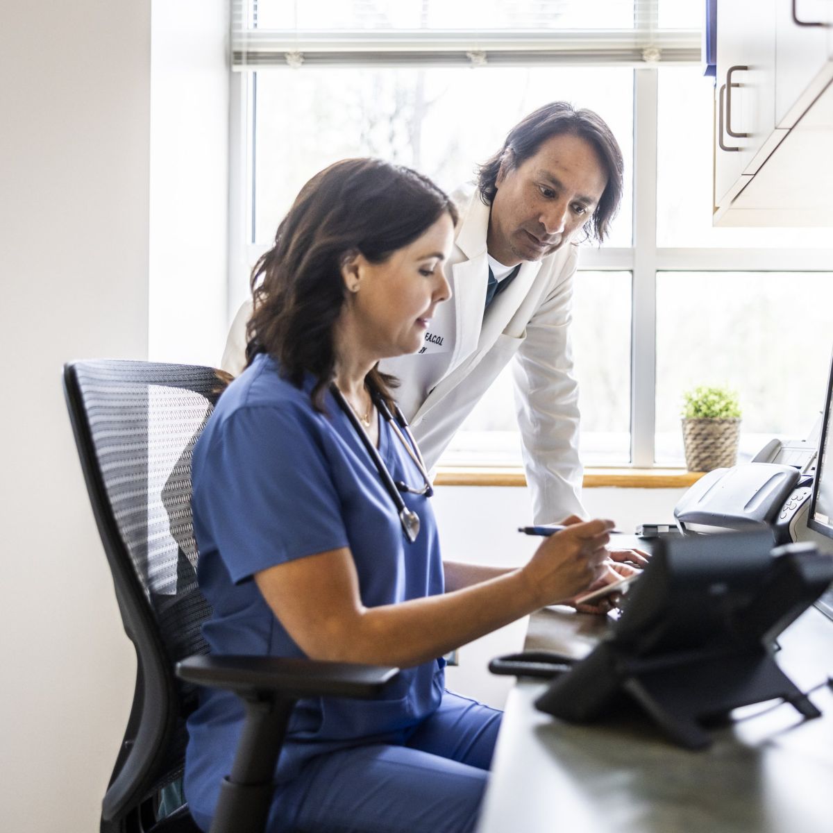 A female nurse and male doctor review digital patient charts together at a workspace.