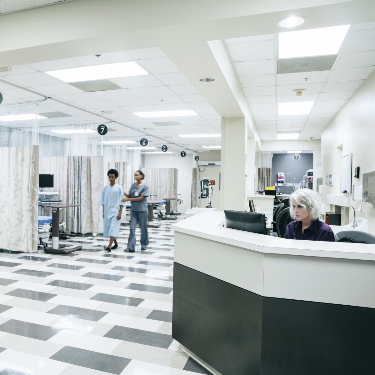 A nurse assists a patient through a modern hospital recovery ward while a staff member works at the nursing station.