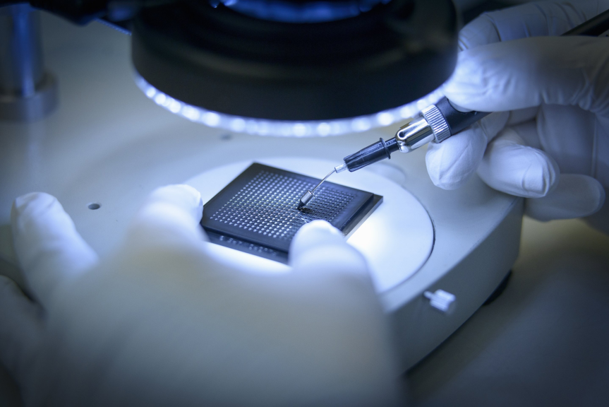 A technician in white gloves uses a probe to inspect a semiconductor chip under a microscope in a laboratory setting.