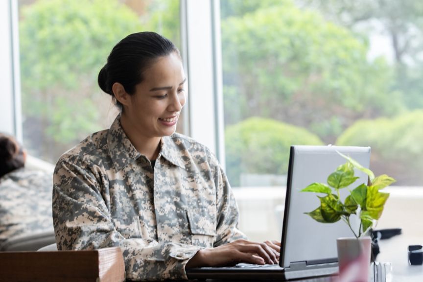 Soldiers in camouflage uniforms using laptops indoors near large windows.