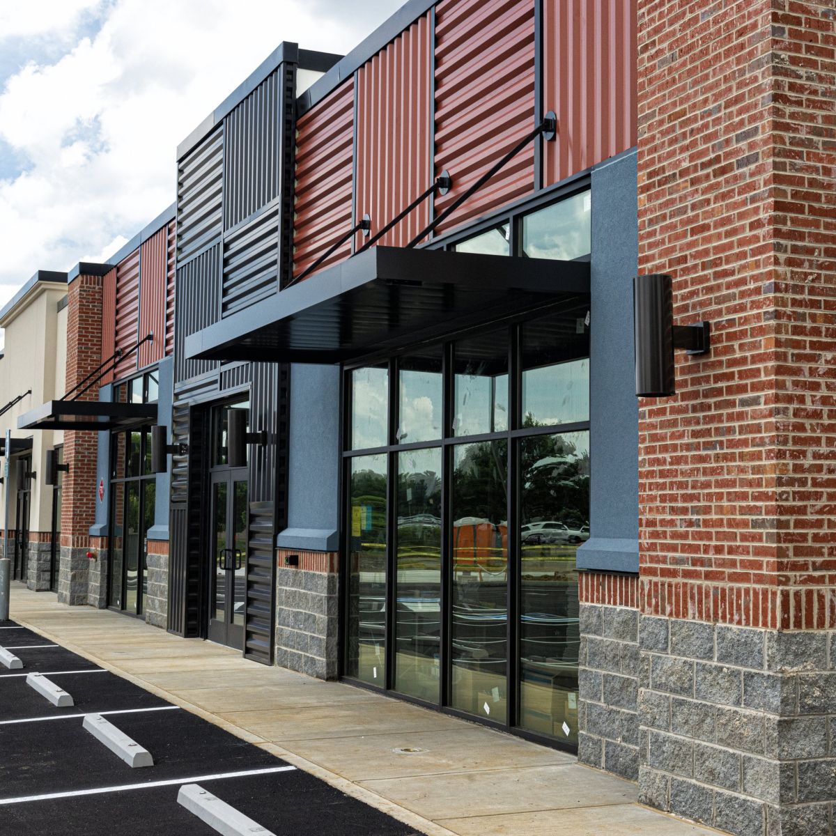 A contemporary retail strip center exterior featuring brick, metal paneling, large glass storefront windows, and an adjacent asphalt parking lot.