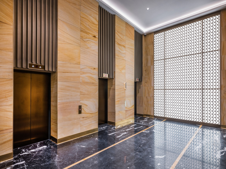 A contemporary corporate lobby featuring sleek bronze elevators, textured stone walls, and a large decorative backlit feature wall over polished marble floors.