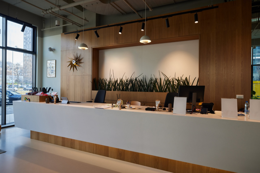 A minimalist corporate reception desk featuring wood paneling, greenery accents, and track lighting in a bright, professional office lobby environment.