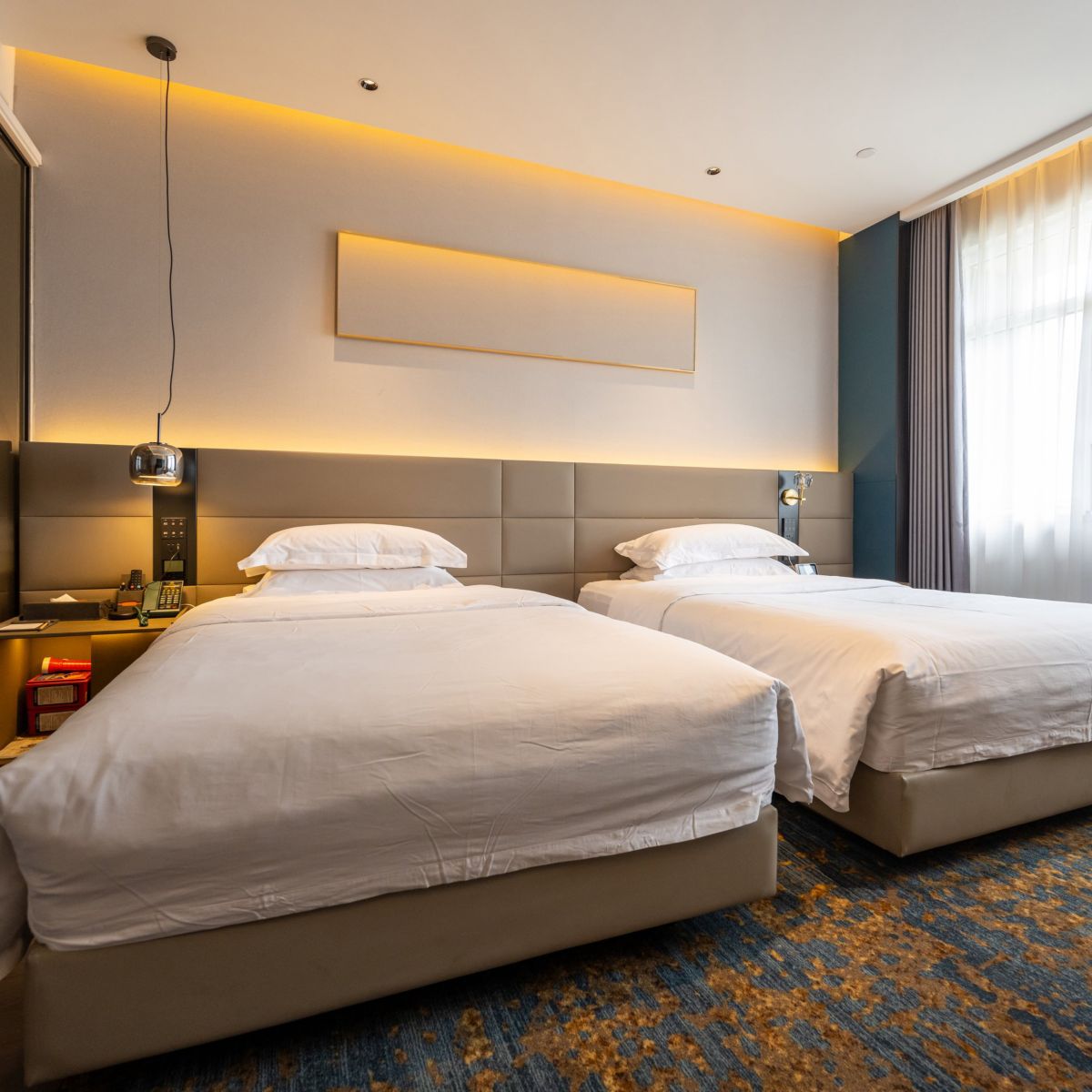 A contemporary hotel room features two neatly made beds, ambient gold cove lighting, and a window with soft sheer curtains over a patterned blue carpet.