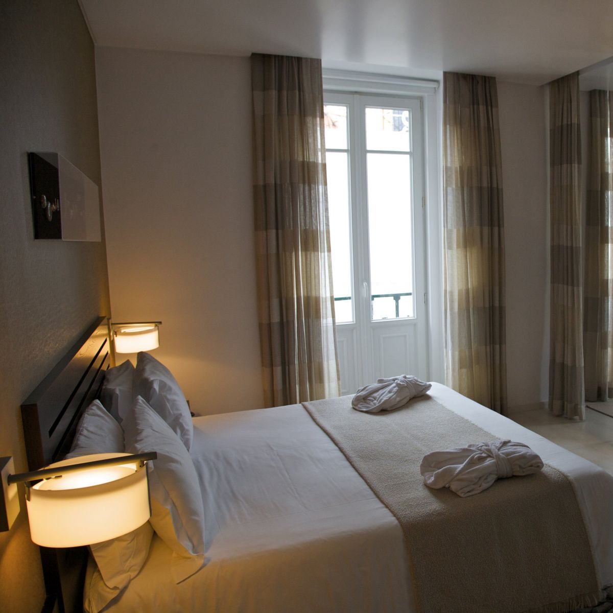 A contemporary, neutrally-toned hotel bedroom featuring a made bed with bathrobes, a wall-mounted television, and a workspace illuminated by warm lighting.