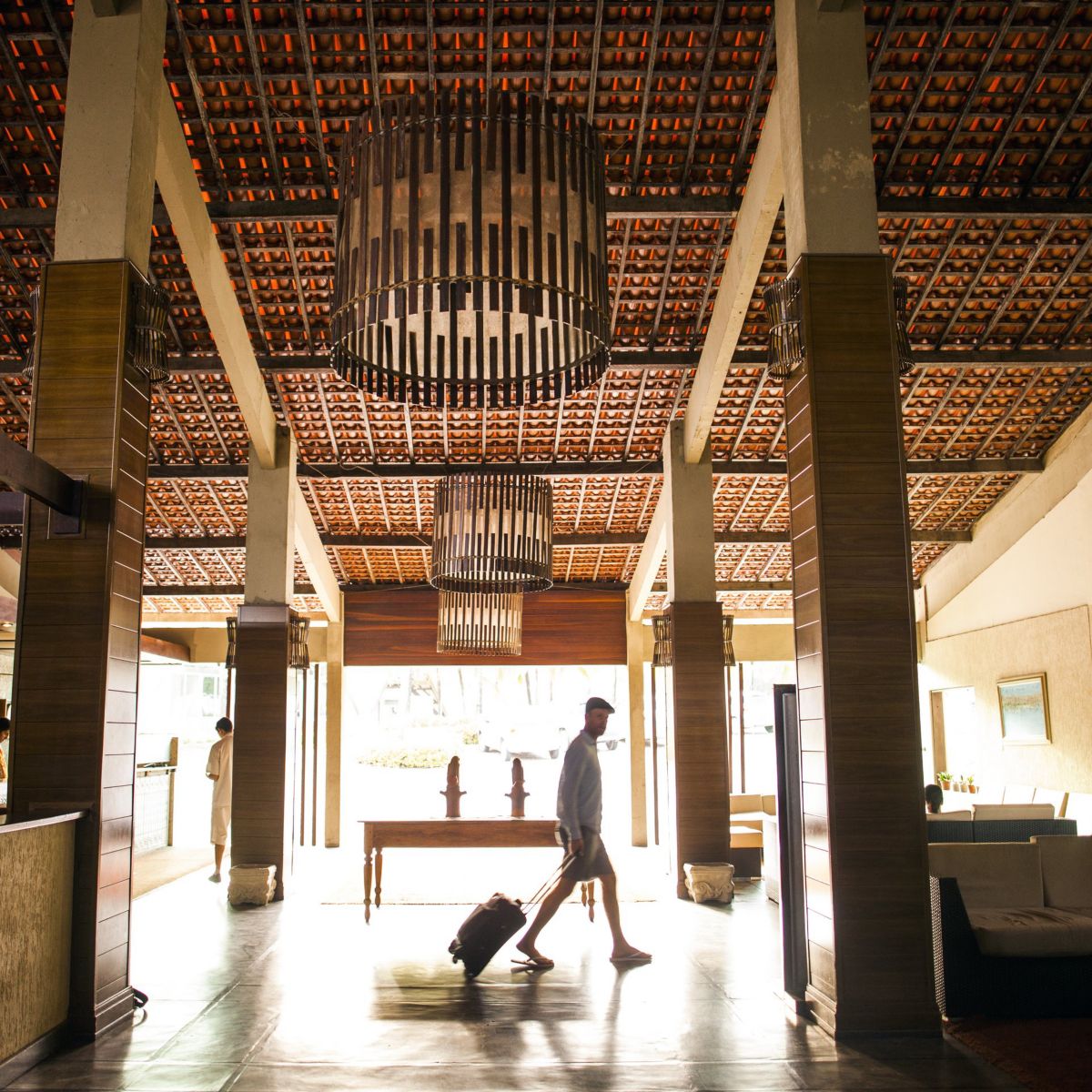 A man with luggage walks through a spacious, high-ceilinged hotel lobby with warm wood accents and contemporary design elements.