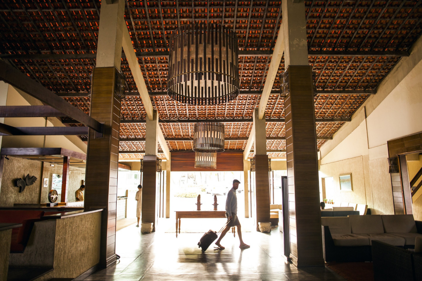 A man with luggage walks through a spacious, high-ceilinged hotel lobby with warm wood accents and contemporary design elements.