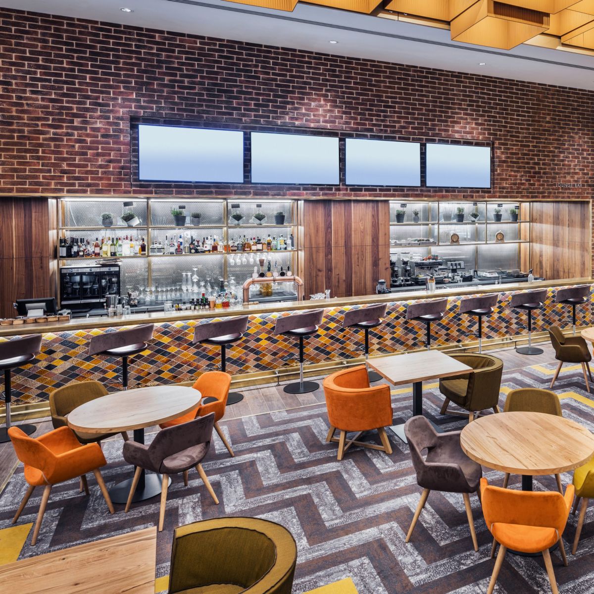 A stylish, contemporary lounge bar featuring a patterned bar front, brick walls, and vibrant, colorful seating arranged across a geometric-patterned carpet.