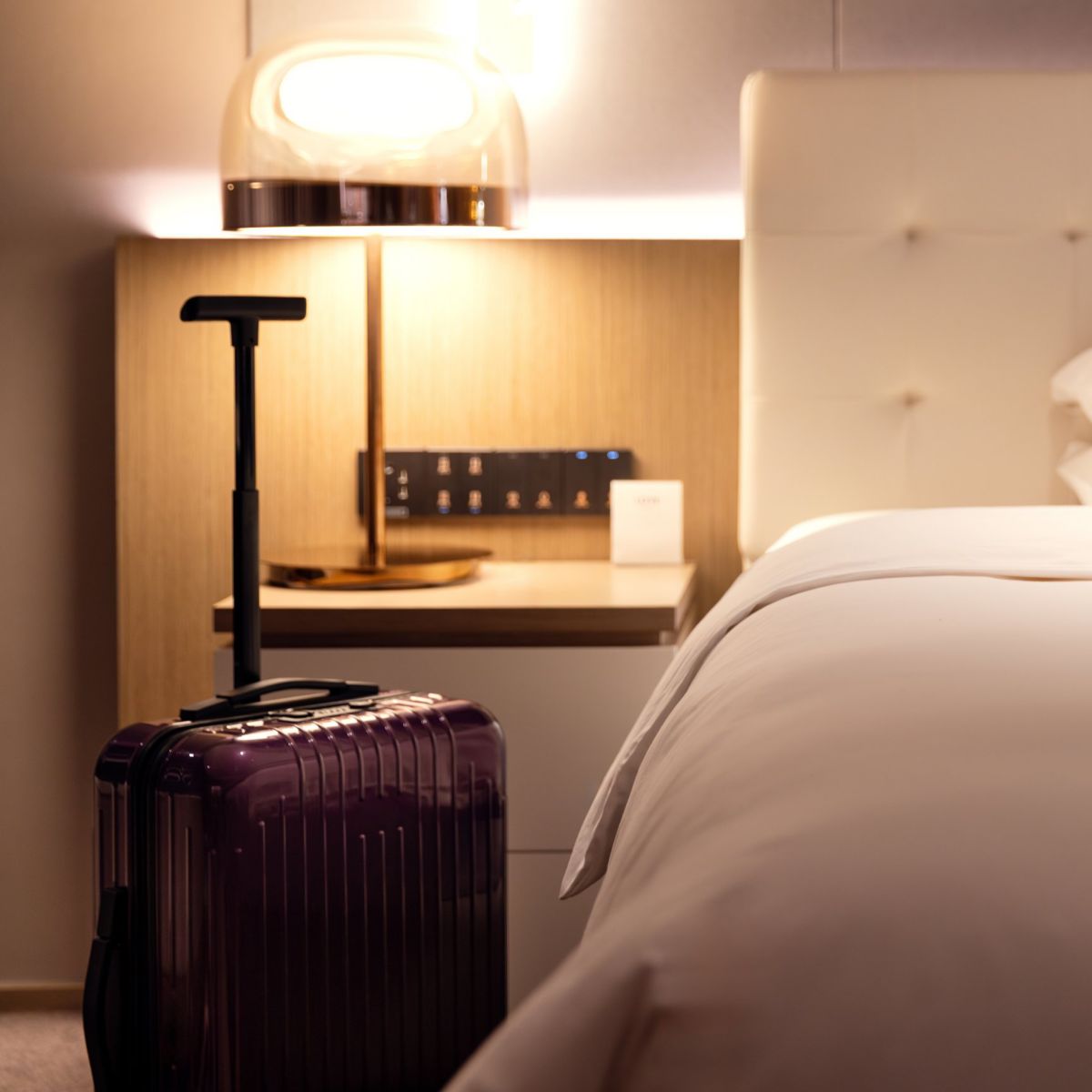 A purple suitcase stands next to a neatly made bed in a contemporary hotel room featuring warm ambient lighting.