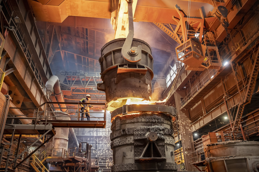 An industrial worker oversees the pouring of molten metal from a crane-suspended ladle into a secondary container at a steel manufacturing plant.